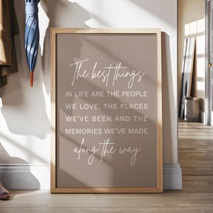 May include: A framed print with the text "The best things in life are the people we love, the places we've been and the memories we've made along the way." The frame is light brown, and the background is a muted brown.