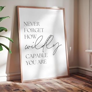 May include: A framed print with the words "NEVER FORGET HOW wildly CAPABLE YOU ARE" in black script and block letters. The frame is a light brown color, and the print is leaning against a wall on a wooden floor.