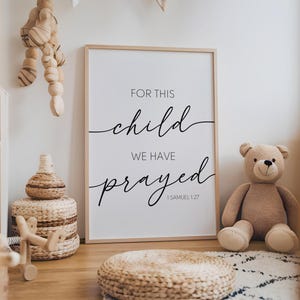 Puede incluir: Una impresión enmarcada blanca con texto negro que dice "Por este niño hemos orado 1 Samuel 1:27". La impresión está colgada en una pared en una guardería con un juguete de mimbre y un oso de peluche.