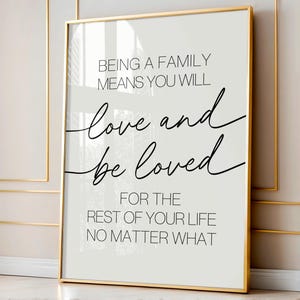 Op de afbeelding: Een wit en grijs printbare wandkunstprint met een gouden lijst. De print bevat een citaat dat luidt: "Een familie zijn betekent dat je zult liefhebben en geliefd zult worden voor de rest van je leven, wat er ook gebeurt."