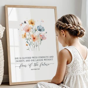 Op de afbeelding: Een aquarelprint met een boeket bloemen in tinten roze, oranje en blauw. De print bevat de quote "Zij is bekleed met kracht en waardigheid, en zij lacht zonder angst voor de toekomst" met de bijbelverwijzing "Spreuken 31:25".