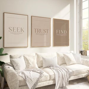 May include: Three framed wall art prints with the words "SEEK", "TRUST", and "FIND" in a neutral color palette. The prints are displayed above a cream-colored sofa with white pillows and a fringed throw blanket. The room has a bright, airy feel.