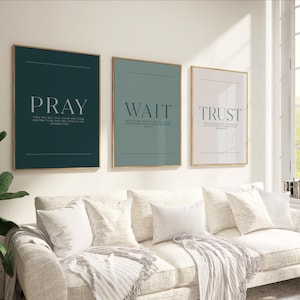 May include: Three framed art prints with the words "PRAY", "WAIT", and "TRUST" in white text. The prints have a minimalist design with a gold frame and a solid color background in teal, sage green, and pale pink. The prints are displayed above a white sofa.