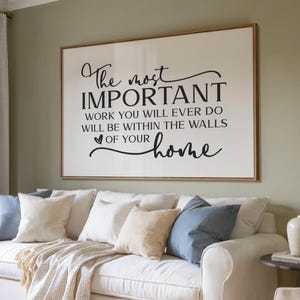 May include: A white framed sign with black text that reads "The most IMPORTANT work you will ever do will be within the walls of your home". The sign is hanging on a green wall above a white couch with pillows.