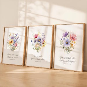 Può includere: Tre stampe artistiche incorniciate con bouquet floreali ad acquerello nei toni del viola, rosa, giallo e blu. Ogni stampa ha una cornice dorata e uno sfondo chiaro con una citazione dei Proverbi. Le stampe sono esposte su una superficie di legno.