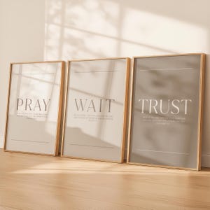 Puede incluir: Tres impresiones artísticas enmarcadas con las palabras "PRAY", "WAIT" y "TRUST" en un diseño minimalista. Cada impresión tiene una paleta de colores neutros con un fondo claro y un marco de madera. Las impresiones se muestran sobre una superficie de madera.