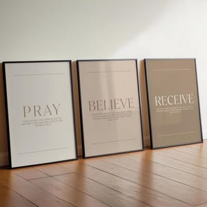 以下が含まれることがあります： 「PRAY」、「BELIEVE」、および「RECEIVE」という言葉が書かれた3つの額入りアートプリント。各プリントは、補完的なフォントのテキストを備えたニュートラルなカラーパレットを備えています。フレームは黒で、プリントは木の床に表示されています。