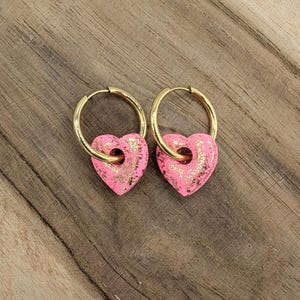 Puede incluir: Un par de pendientes de aro dorados con dijes de corazón rosa. Los corazones tienen un acabado de brillo dorado.