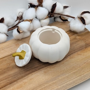 Peut inclure: Un récipient blanc en céramique en forme de citrouille avec un couvercle et une tige dorée. Le couvercle est à côté du récipient. Le récipient est sur une surface en bois, avec des branches de coton en arrière-plan. Le récipient est probablement à usage décoratif.