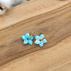 Könnte beinhalten: Ein Paar blütenförmige Ohrringe. Jeder Ohrring hat ein hellblaues und weißes Marmormuster mit einem goldfarbenen Reifen. Die Ohrringe sind vor einem Holzgrund, mit Baumwollzweigen im Hintergrund.