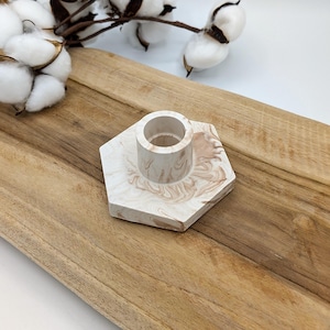 Peut inclure: Un bougeoir hexagonal en marbre blanc et brun avec une ouverture ronde pour une bougie. Le bougeoir est posé sur une surface en bois.