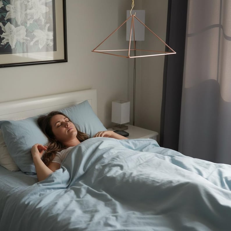 May include: A copper-colored metal pyramid frame suspended from the ceiling by a thin chain. The geometric structure features a square base and four triangular sides meeting at a single top point, hanging above a bed with light blue bedding.