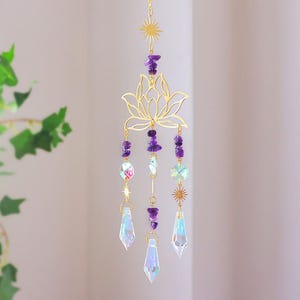 May include: A gold-colored lotus flower suncatcher with hanging amethyst chips and iridescent crystal prisms. The suncatcher is suspended by a delicate chain and features small sunburst accents. The overall design is elegant and decorative.