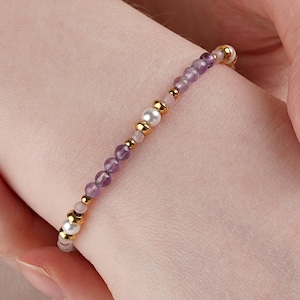 Op de afbeelding: Een delicate armband met amethistkralen, kleine roze kralen en gouden accenten. De armband bevat ook een grote witte parel. De armband is ontworpen om om de pols te worden gedragen.