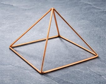 Puede incluir: Una escultura piramidal geométrica construida con alambre metálico fino de color oro rosa. La estructura presenta una base cuadrada con cuatro lados triangulares que se unen en un solo vértice, creando un diseño de marco abierto y hueco.