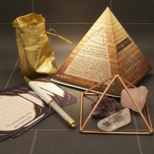 Puede incluir: Un surtido de artículos espirituales, que incluyen una bolsa dorada con cordón, una caja en forma de pirámide, una pirámide de cobre, cristales y un bolígrafo. La caja dice "Crystal Jewels and Pyramid Energizer". El bolígrafo tiene un barril transparente lleno de cristales.