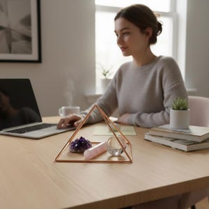 May include: A copper-colored metal pyramid frame containing a purple amethyst crystal cluster, a smooth cylindrical rose quartz stone, and a clear glass sphere, all arranged together on a light-colored wooden surface.