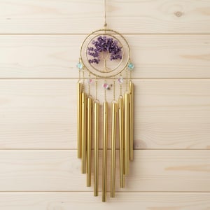 May include: A gold-colored wind chime with a circular design featuring a tree made of purple stones. The chime has multiple gold tubes hanging below, with colorful beads and chains.