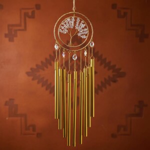 Puede incluir: Un carill&oacute;n de viento dorado con un dise&ntilde;o de &aacute;rbol de la vida. El &aacute;rbol est&aacute; dentro de un marco circular, adornado con cristales transparentes. Debajo, m&uacute;ltiples tubos dorados cuelgan de cadenas. El fondo es marr&oacute;n c&aacute;lido con patrones geom&eacute;tricos.