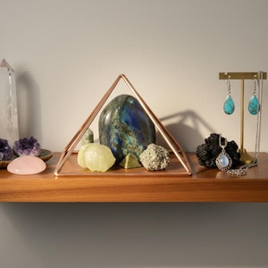 May include: A copper-colored wire pyramid frame containing a large blue and green stone, a pale yellow stone, a small green pyramid, and a textured metallic stone. Nearby are a pink stone, a purple geode, and a necklace and earring set.