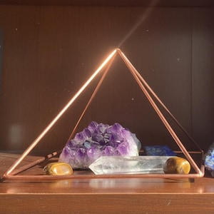 May include: A copper wire pyramid frame enclosing a collection of crystals. Inside the frame sits a purple amethyst cluster, a clear quartz point, two smooth brown tiger's eye stones, and a small piece of blue sodalite.