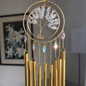Puede incluir: Carrill&oacute;n de viento dorado con dise&ntilde;o de &aacute;rbol de la vida. El &aacute;rbol est&aacute; hecho con cristales transparentes y alambre, dentro de un marco de doble anillo. Cuelgan debajo campanillas doradas y detalles de cristal iridiscente, creando una pieza decorativa.