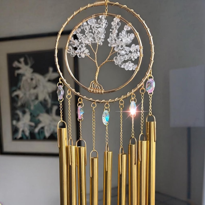Puede incluir: Carrill&oacute;n de viento dorado con dise&ntilde;o de &aacute;rbol de la vida. El &aacute;rbol est&aacute; hecho con cristales transparentes y alambre, dentro de un marco de doble anillo. Cuelgan debajo campanillas doradas y detalles de cristal iridiscente, creando una pieza decorativa.