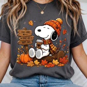 Könnte beinhalten: Ein anthrazitfarbenes T-Shirt mit einer Grafik eines weißen Hundes, der eine orangefarbene Strickmütze und einen Schal trägt und eine Kaffeetasse hält. Das Design zeigt ein Holzschild mit dem Text COZY VIBES, CRISP AIR, HAPPY DAYS, einen orangefarbenen Kürbis und Herbstblätter.
