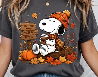 May include: A charcoal gray t-shirt featuring a graphic of a white dog wearing an orange knit hat and scarf, holding a coffee cup. The design includes a wooden sign with the text COZY VIBES, CRISP AIR, HAPPY DAYS, an orange pumpkin, and autumn leaves.