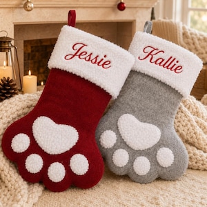 May include: Two paw-shaped Christmas stockings, one red and one gray, featuring white sherpa cuffs embroidered with the names Jessie and Kallie in red script. Each stocking has a large white sherpa heart-shaped pad and four smaller toe pads.
