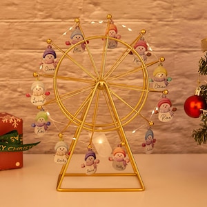May include: A gold-colored Ferris wheel Christmas ornament with small snowman figures hanging from the wheel. Each snowman has a different colored hat and scarf. The names of the people are written on the snowmen. A string of lights is wrapped around the wheel.