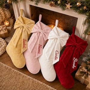 Op de afbeelding: Vier kerstsokken van fluweel in mosterdgeel, lichtroze, wit en donkerrood. Elke sok heeft een grote decoratieve strik aan de bovenkant en een ophanglus. De namen Isabel, Naomi, Carlie en Brooke zijn in het rood of wit geborduurd.