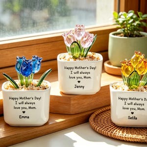 Peut inclure: Trois petits pots carrés blancs avec des fleurs de tulipes en verre bleues, roses et jaunes. Chaque pot porte l'inscription "Joyeuse fête des mères ! Je t'aimerai toujours, maman." et un nom. Une petite plante verte est en arrière-plan.