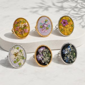 May include: Six oval rings featuring pressed flowers encased in resin. The collection includes rings with yellow, lavender, and black backgrounds, each decorated with various small colorful blossoms, green foliage, and tiny white pearl accents.