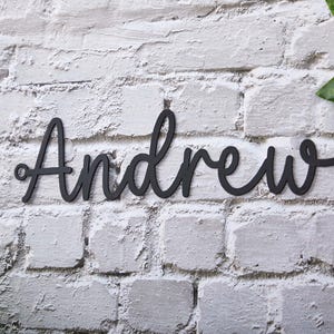 Puede incluir: La palabra "Andrew" en negro, escrita en cursiva, está montada en una pared de ladrillos blancos. Las letras están conectadas y tienen una apariencia suave y elegante. La pared tiene un aspecto texturizado y envejecido.
