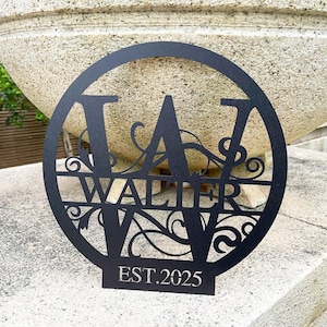 May include: Black metal sign featuring the name "Walter" and initials "W A V" within a circle. The sign includes the text "EST. 2025". The design incorporates decorative flourishes. A decorative item.