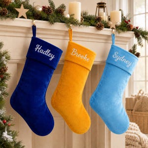 Könnte beinhalten: Drei Weihnachtssocken aus Samt hängen in einer Reihe. Die linke Socke ist dunkelblau mit dem in Weiß gestickten Namen Hadley. Die mittlere Socke ist senfgelb mit dem in Weiß gestickten Namen Brooke. Die rechte Socke ist hellblau mit dem Namen Sydney.