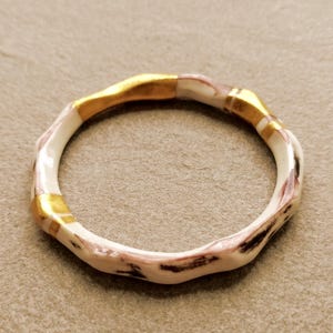 Könnte beinhalten: Ein kreisförmiger Ring mit einer wellenförmigen, organischen Form. Das Band besteht aus einem glatten, cremeweißen Material mit dunkelbraunen, holzähnlichen Maserungen und ist mit drei markanten, polierten goldfarbenen Metallsegmenten verziert.