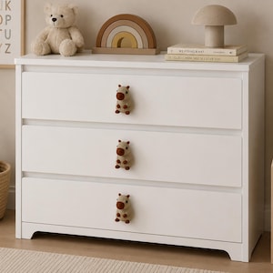 May include: A white three-drawer chest of drawers with animal-shaped knobs. The chest is adorned with a teddy bear, a wooden rainbow, and a mushroom lamp. An alphabet poster and a woven basket with a stuffed bunny are also visible.