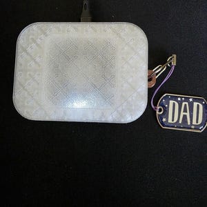 May include: A white, square wireless charger with a textured pattern and a connected black cable. A small, blue and white tag with the word "DAD" is attached with a purple string and metal clasp.