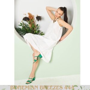 May include: A woman wearing a white, sleeveless dress with a V-neckline and a tiered hem. She is wearing green heeled sandals. The background is a light green wall with a white circular cutout. The text "BOHEMIAN BREEZES 444" is at the bottom.