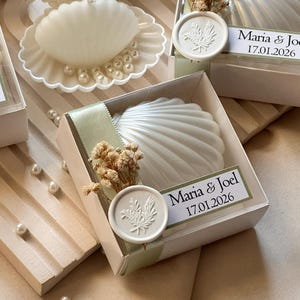 Puede incluir: Jabones blancos en forma de concha en cajas transparentes, algunos con perlas decorativas. Cada caja está adornada con una cinta, flores secas y una etiqueta que dice "Maria & Joel 17.01.2026".