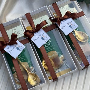 May include: Three rectangular white gift boxes containing vanilla latte coffee packets and gold-toned decorative spoons. Each box is tied with a brown satin ribbon and features a small white tag with the text With Love and a couple illustration.