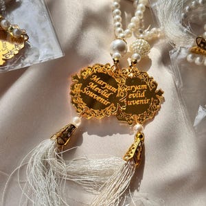 May include: Gold-colored pendants with the text "Maryam Mevlid Souvenir" are attached to a pearl necklace. The pendants have a decorative border and are paired with white tassels. Several similar items are visible.