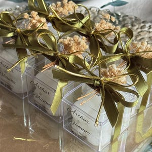 May include: Clear square boxes filled with white items, adorned with olive green satin ribbons and small dried flower bouquets. The boxes are printed with the text "Arthur & Rihanna" and a date. They are arranged on a clear, reflective surface.