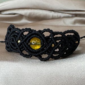 Peut inclure: Un bracelet en macramé noir orné d'une pierre précieuse jaune centrale et de petites perles dorées. Le bracelet est finement noué et repose sur une surface satinée, mettant en valeur le savoir-faire de la bijouterie.