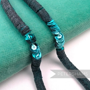 May include: Close-up of two strands of dark gray sequin trim with teal accents. The sequins are circular and reflect light, creating a shimmering effect. The trim is set against a teal background, with the word "PETERSHAMS" in white.