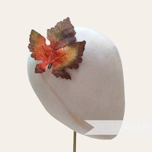 May include: A white foam hat form with a single brown, orange, and red leaf attached to the side. The hat form is on a gold stand. The text "PETERSHAMS" is visible on the bottom right side of the hat form.