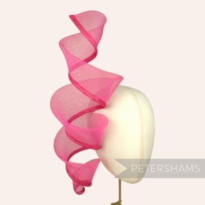 以下が含まれることがあります： 鮮やかなピンク色のヘッドピース。渦巻くリボンのようなデザイン。マネキンの頭に取り付けられています。リボンにはキラキラしたトリムが付いています。「PETERSHAMS」の文字がマネキンの頭に印刷されています。