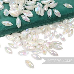 May include: A collection of iridescent, oval-shaped beads scattered on a green velvet surface. The beads have a pearlescent sheen, reflecting a spectrum of colors. The image includes the text "PETERSHAMS".